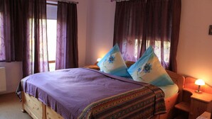 Quarto com cama de madeira, roupa de cama roxa, duas almofadas com estampas florais, abajur e janela com cortinas.