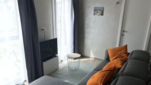 A modern living room with a grey sofa, orange cushions, a small round table, and a television on a white cabinet. - Lovely vacation home, De Haan, 6 pers., completely renovated, child-friendly (De Haan)