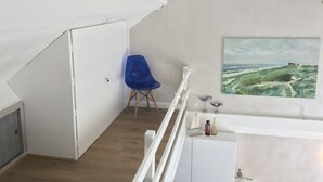 A room with a sloped ceiling, a blue chair, a painting of a landscape, and a white cabinet with decorative items. - Lovely vacation home, De Haan, 6 pers., completely renovated, child-friendly (De Haan)