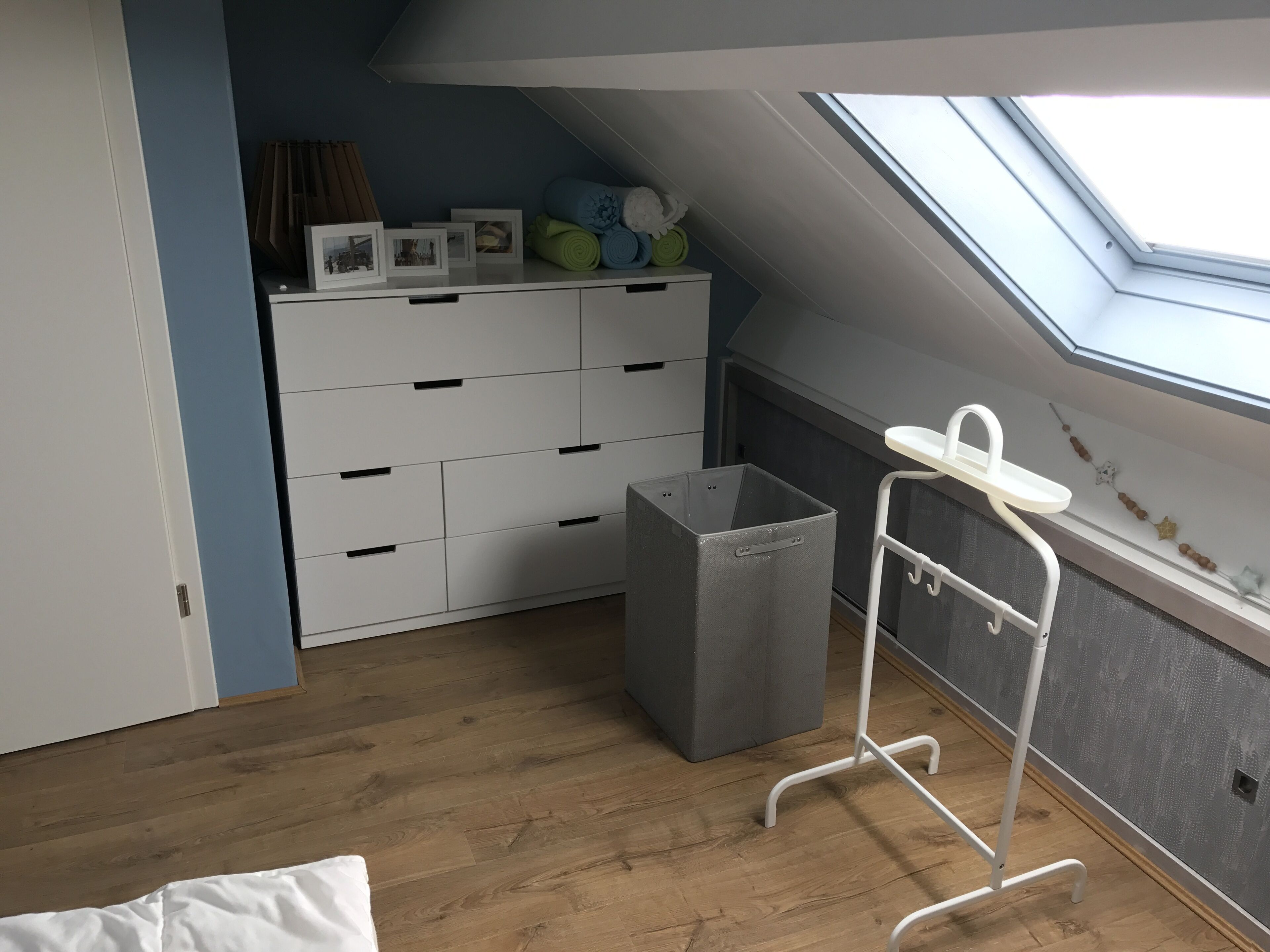 Una habitación con tragaluz, un mueble de almacenamiento blanco, un cubo de basura gris y una estantería metálica blanca cerca de una ventana.