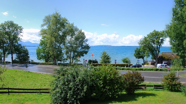 A coastal road with a view of the sea and mountains. There are trees, a grassy area, and a building with a sign.