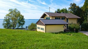 A two-story house with a sloped roof, a visible chimney, and a small porch. The house is situated on a grassy hill with a view of a lake.