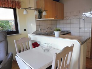 Uma cozinha compacta com bancada branca, pia e fogão. Há uma janela com persianas, uma cadeira e uma mesa com uma cesta vermelha.