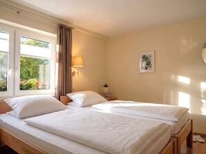 Cama doble con ropa de cama blanca en una habitación con paredes color beige, una ventana con cortinas y una lámpara fijada a la pared.