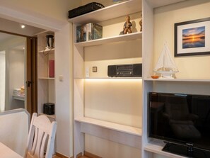A modern living room with a flat-screen TV, a bookshelf with books and decorative items, a small table with a chair, and a view of the outside through a window.