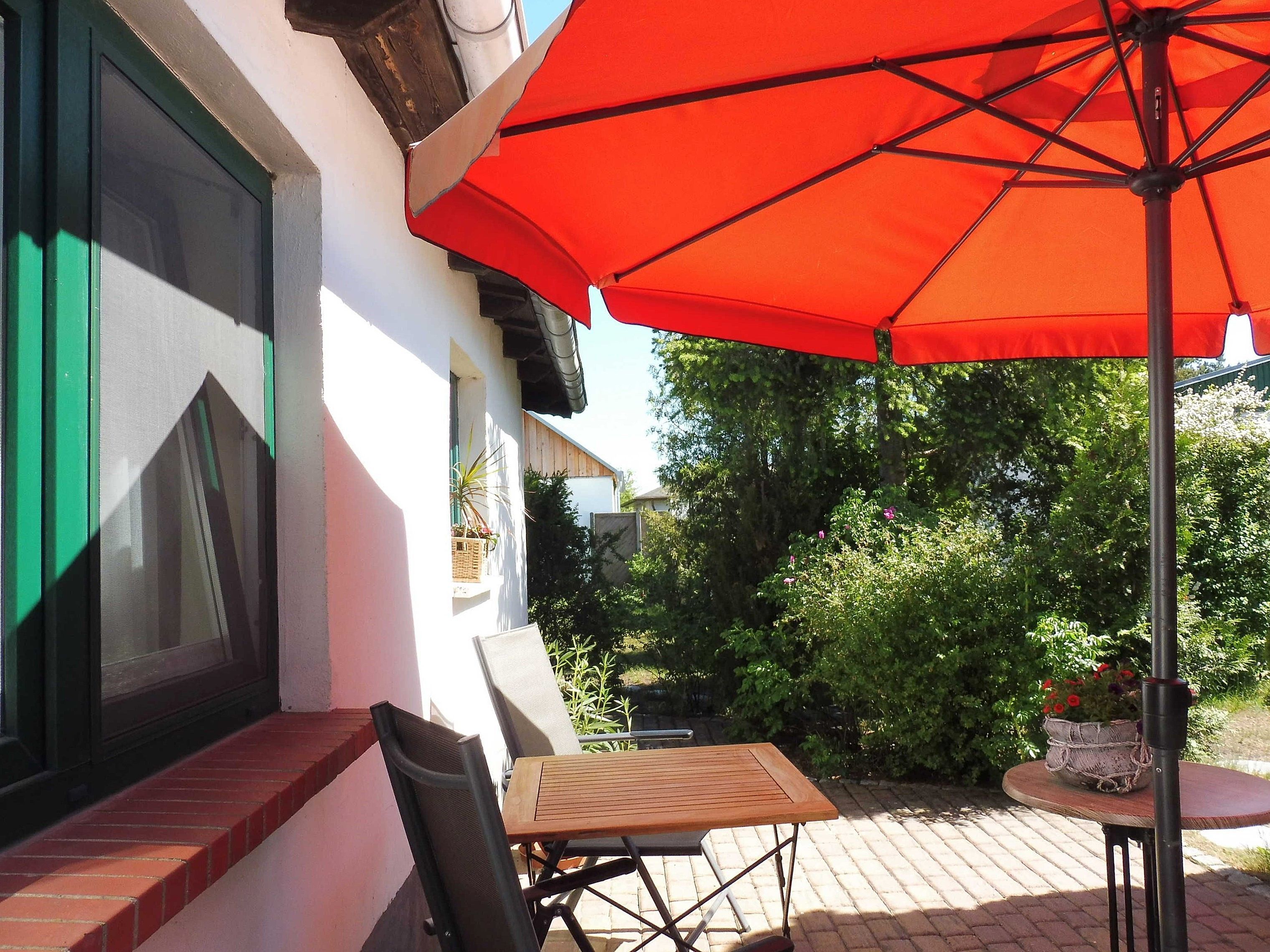 Een terras met een tafel, stoelen, een rode parasol en een plant in een pot.