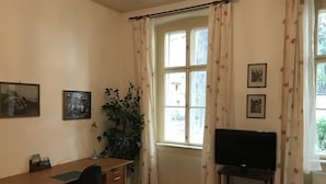 Ein Zimmer mit Schreibtisch aus Holz, einem schwarzen Bürostuhl, einem Fernseher auf einem Beistelltisch, einer Topfpflanze und einem Fenster mit blumigen Vorhängen.