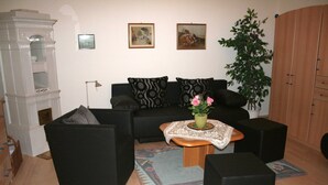 Ein Wohnzimmer mit einer schwarzen Couch, zwei schwarzen Sesseln, einem Holztisch mit Blumenvase, einem gemusterten Teppich und einer großen Grünpflanze.