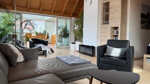 A modern living room with a wooden ceiling, a black leather sofa, a black armchair, and a glass door leading to an outdoor area. - Apartment 90 square meters, open living area with WiGarten, 2 bedrooms, max 5 p. (Kressbronn)