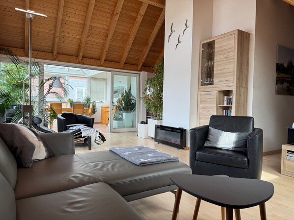 A modern living room with a wooden ceiling, a black leather sofa, a black armchair, and a glass door leading to an outdoor area.