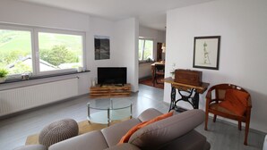 Una sala de estar con un sofá, una mesa de centro de cristal, una televisión y un piano. Se ven ventanas con vistas a zonas verdes.