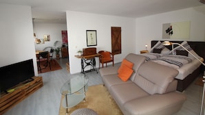 Una sala moderna con una mesa de centro de vidrio, un sofá con cojines naranjas, un televisor sobre un soporte de madera, y una zona de comedor al fondo.