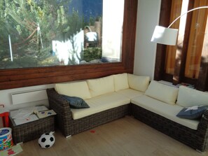 A room with a wicker sofa, a window with a view of greenery, a lamp, and a basket with items on the floor. - First class 2 room apartment, 2-4 people, close to the center of congress (Graubünden)