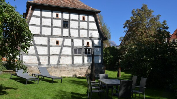 A traditional half-timbered house with a stone foundation, a lawn with outdoor furniture, and a clear blue sky.