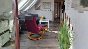 Ein modernes Wohnzimmer mit einem roten Sessel, einem grauen Sofa und einem weißen Couchtisch. Ein Fenster mit weißen Vorhängen, ein Heizkörper und ein gerahmtes Bild sind an der Wand zu sehen.