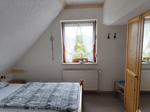 Ein Schlafzimmer mit schrägverdeckter Decke, einem Fenster mit Spitzenvorhängen, einem Heizkörper, einem Bett mit gemustertem Überwurf, einem Holzstuhl und einem Holzschrank.