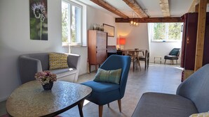 Ein Wohnzimmer mit einer Couch, einem Sessel und einem kleinen Tisch mit Blumen. Es gibt einen Holzschrank, einen Esstisch mit Stühlen und ein Fenster mit Blick auf Grünflächen.