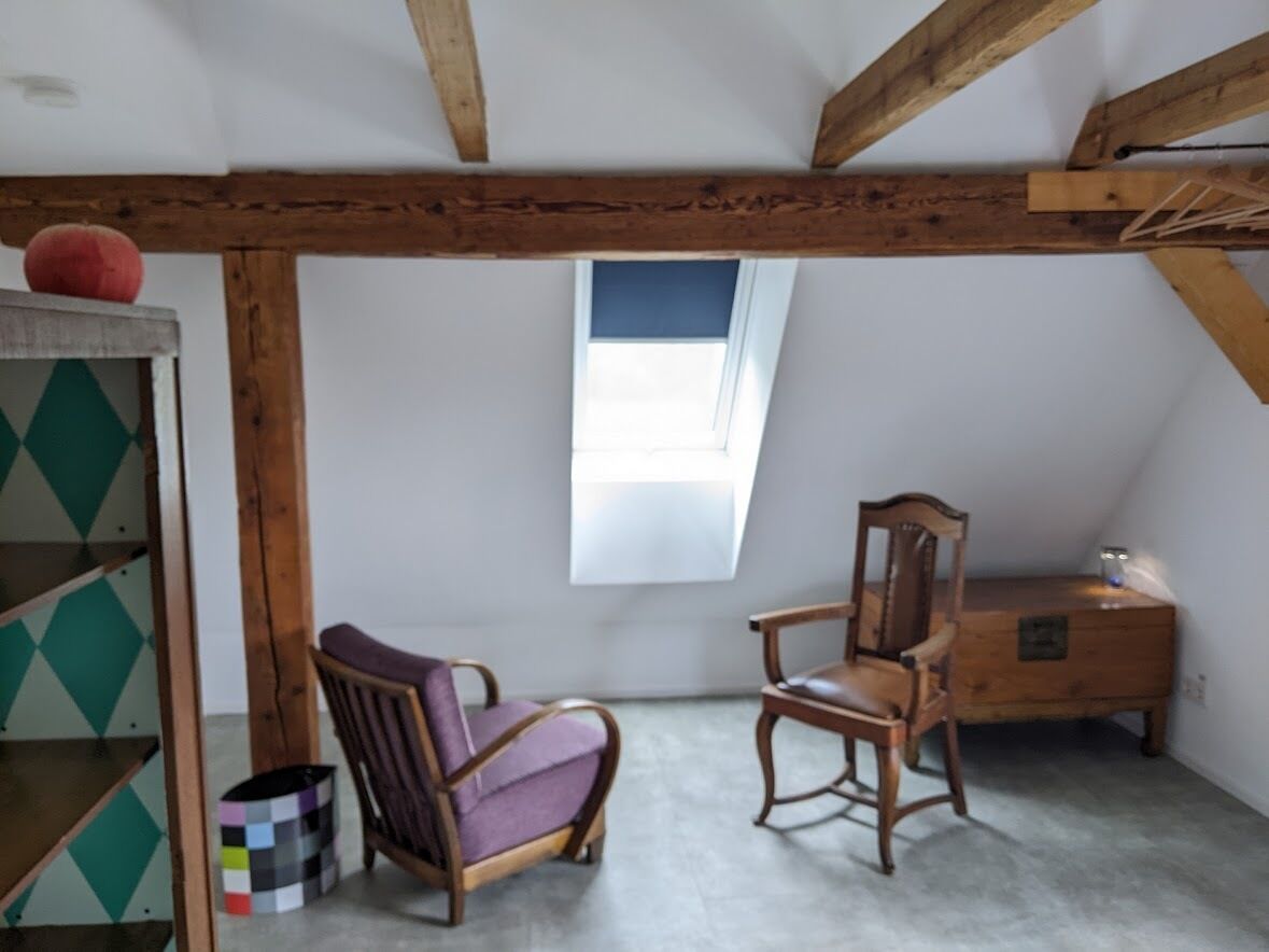 A room with a sloped ceiling, wooden beams, a window, a chair, a wooden dresser, and a shelf with a geometric-patterned box.