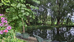 Eine Holzterrasse mit grünen Metallstühlen mit Blick auf einen ruhigen See, umgeben von üppiger Vegetation und blühenden Pflanzen.