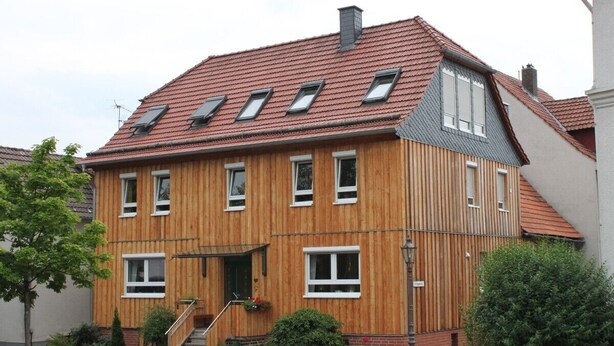 Ein zweistöckiges Holzhaus mit geneigtem Dach, mehreren Fenstern und einer kleinen Veranda.