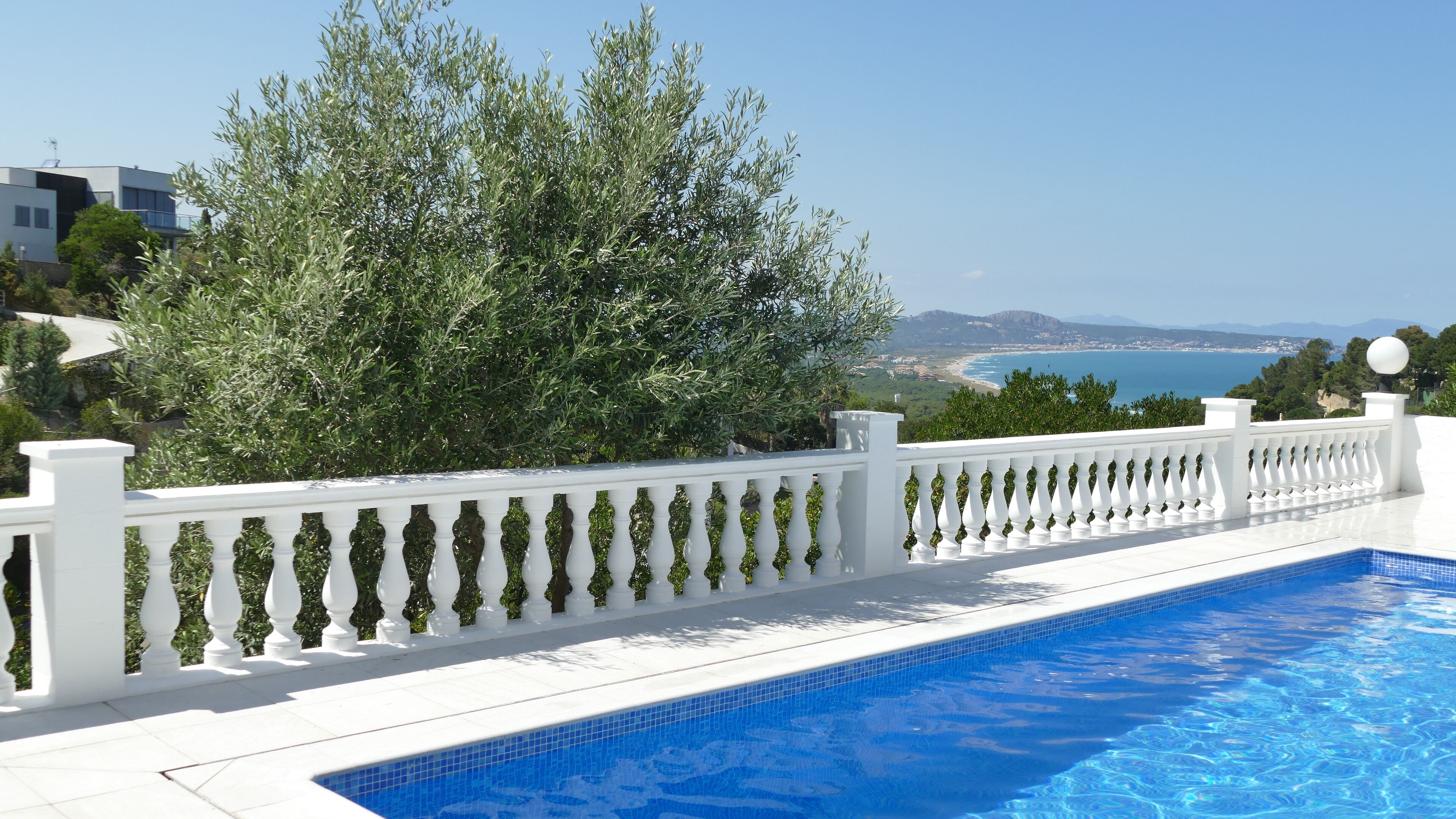 A swimming pool with a white balustrade, surrounded by greenery and overlooking a coastal view.