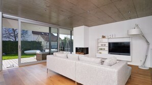 A modern living room with a large sectional sofa, a flat-screen TV mounted on the wall, and a sliding glass door leading to an outdoor patio. - LOFT Rebhalde in Nonnenhorn on Lake Constance offers relaxation and comfort at the same time (Nonnenhorn)