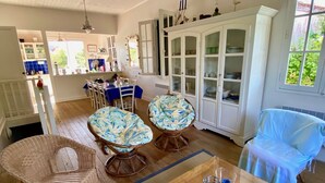 A spacious dining area with a wooden table, two wicker chairs, and a blue-covered chair. A white cabinet with glass doors is visible in the background. - Fantastic house with idyllic garden in the heart of Cap Ferret, very central (Lège-Cap-Ferret)