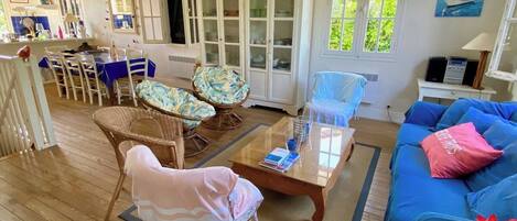 Un salon avec un canapé bleu, des fauteuils en osier, une table basse en verre et un meuble avec des portes en verre.