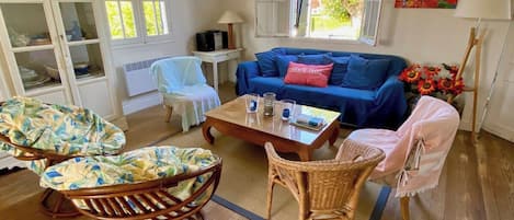 Ein Zimmer mit einem blauen Sofa, Korbstühlen, einem Holztisch und Blick auf Grün.