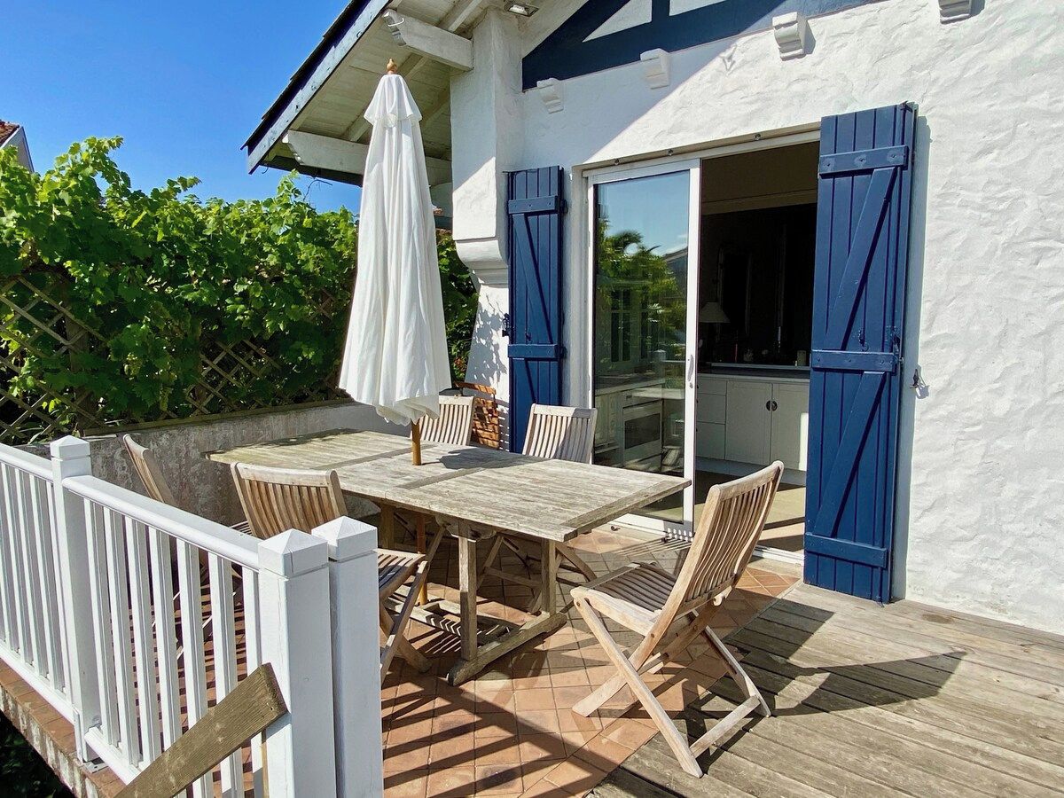 Une terrasse extérieure avec une table et des chaises en bois, un parasol blanc et une vue sur l’intérieur de la maison à travers une porte vitrée.