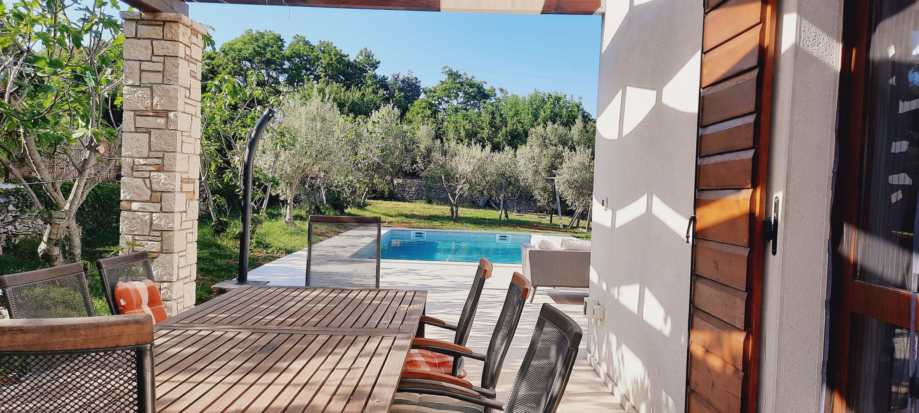 An outdoor patio with a wooden table and chairs, a stone pillar, and a view of a swimming pool and trees.