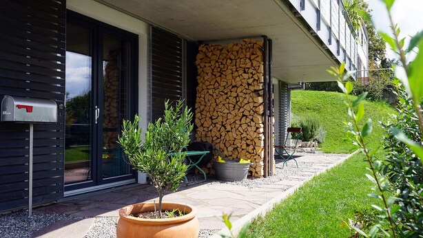 A modern building with a glass door entrance, a small potted plant, a stack of firewood, and a mailbox. - FeWo Betz Bodensee in Überlingen (Überlingen)