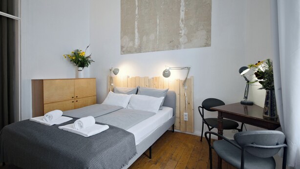 A modern hotel room with a wooden headboard, a large bed with white linens, a desk with a chair, and a wall-mounted lamp. - Renovated old apartment with shower room, separate eat-in kitchen and washing machine (Berlin)