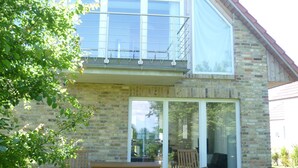 A two-story stone house with a balcony, a patio with a table and chairs, and a large window. - Haus Mirabelle: Baltic Sea view through trees close to the beach (5 min walk) (Kalkhorst)