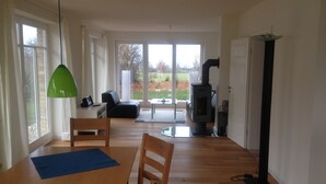 A modern living room with a wooden dining table, chairs, a green pendant light, a black sofa, and a large window with a view of the outdoors. - Haus Mirabelle: Baltic Sea view through trees close to the beach (5 min walk) (Kalkhorst)