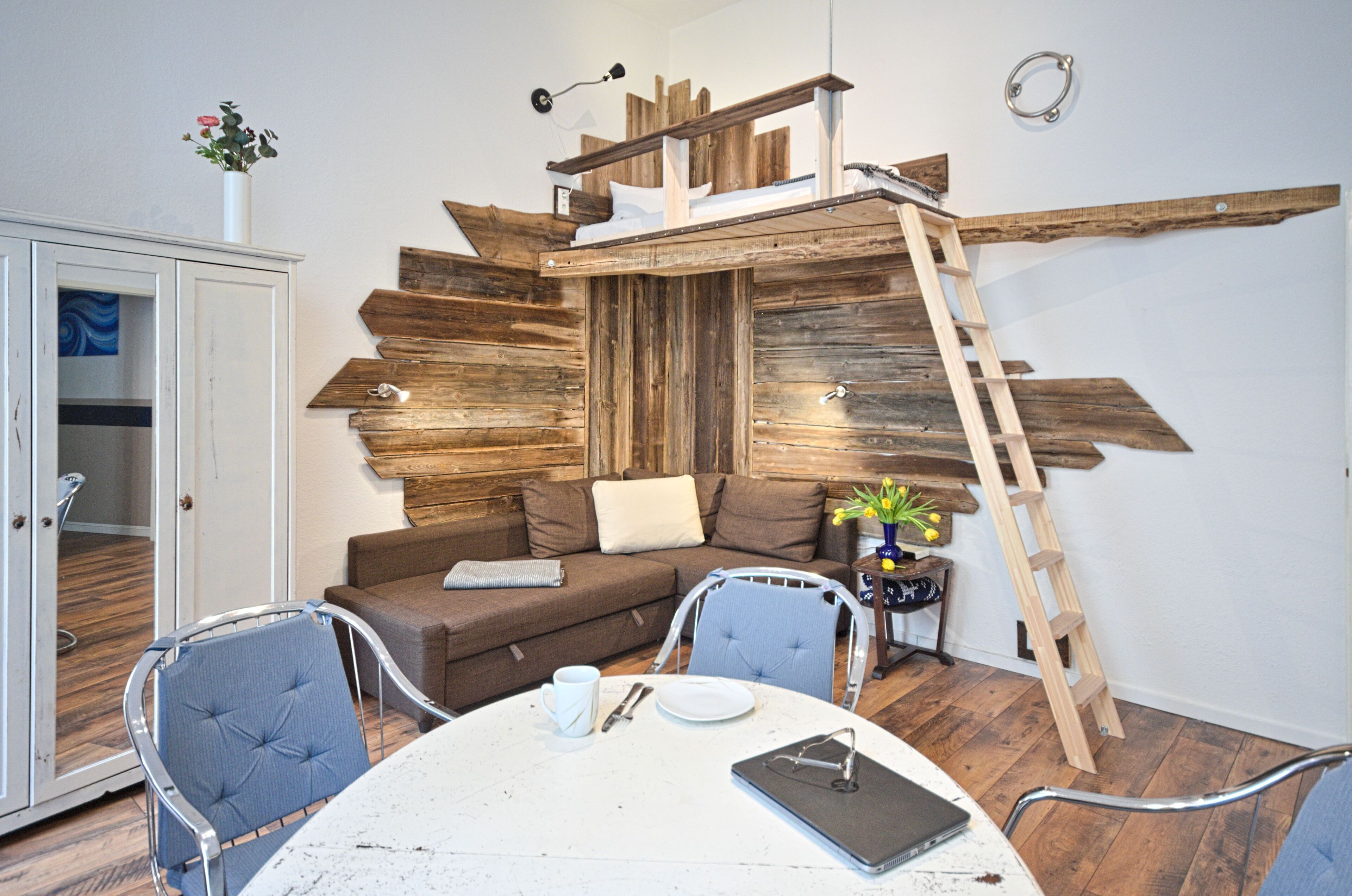 Ein gemütliches Wohnzimmer mit einer Wand aus Holzpaneelen, einem Schlafplatz in der Erhöhung, einem Esstisch mit Stühlen und einem Schrank.