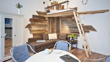 Ein gemütliches Wohnzimmer mit einer Wand aus Holzpaneelen, einem Schlafplatz in der Erhöhung, einem Esstisch mit Stühlen und einem Schrank.