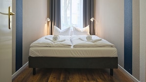 Een netjes opgemaakt bed met witte lakens, een donker hoofdbord en een raam met gordijnen.