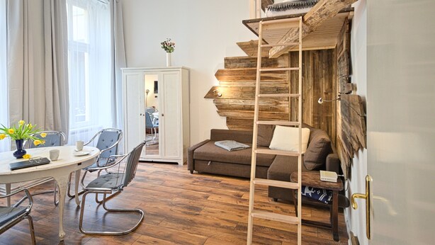 Een kamer met een houten wenteltrap, een bank, een eettafel met stoelen en uitzicht op een stadsgezicht door een raam.