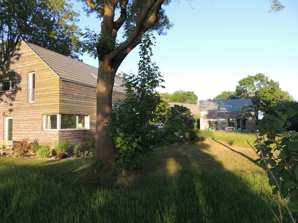 A residential property with a large tree in the front yard, a wooden house with a sloped roof, and a smaller building with a flat roof in the background.
