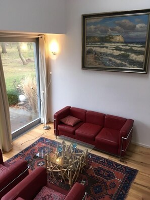 Un salon avec un canapé rouge, une table basse en verre, un tapis à motifs, un tableau représentant un paysage marin et une vue à travers une porte vitrée.