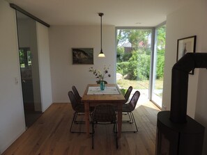 Un coin repas avec une table en bois, des chaises en osier, un vase de fleurs, une suspension et une vue sur la verdure à l’extérieur.