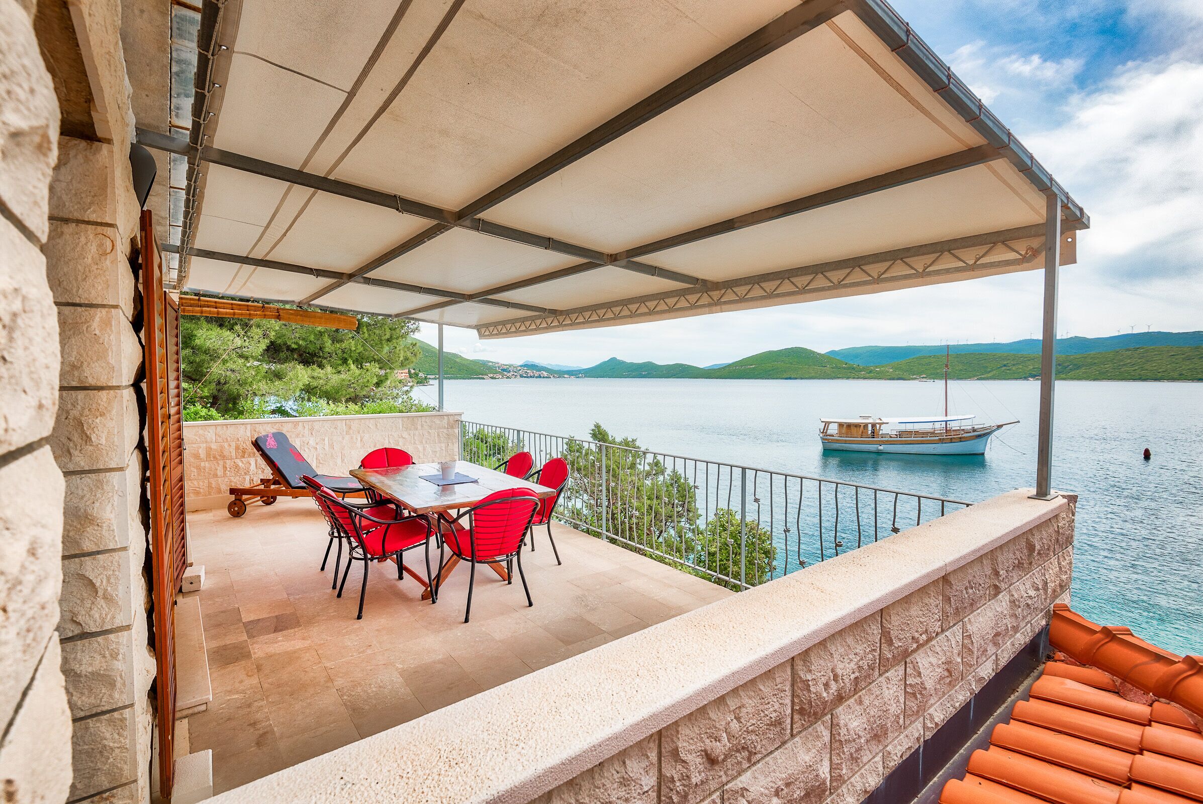 A covered outdoor dining area with a table and chairs, overlooking a body of water and hills.