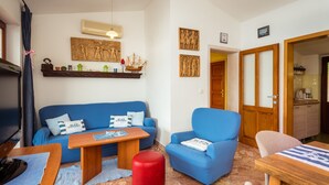 A living room with a blue sofa, a wooden coffee table, a red ottoman, and a blue armchair. There is a television on the left and a wooden door leading to the kitchen. - Nice villa direct on sea,surrounded with lot of mediterranean plants,two minutwe (Klek)