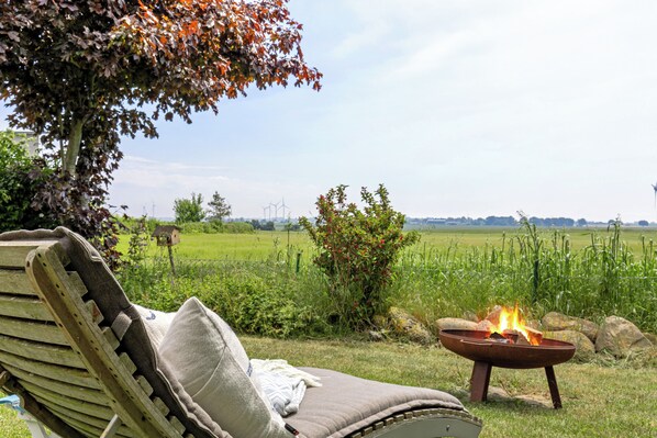 A lounge chair with a cushion, a fire pit with flames, and a view of a green field and wind turbines in the background. - On the wildly romantic natural beach, enchanting view, cozy with stove, sauna (Schleswig Holstein)