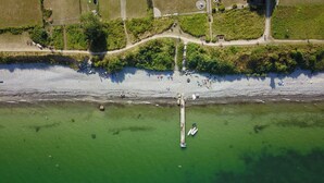 An aerial view of a beach with a pier extending into the water, surrounded by greenery and a path along the shore.