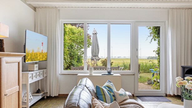 A living room with a television, a sofa, a window with a view of a field and a lighthouse, and a door leading outside.