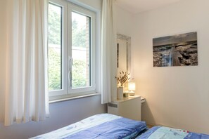 A bedroom with a bed, a window with curtains, a bedside table with a lamp, and a wall-mounted mirror. - On the wildly romantic natural beach, enchanting view, cozy with stove, sauna (Schleswig Holstein)