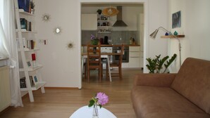 Un salon avec un canapé marron, une table basse blanche ornée d’un vase de fleurs, une bibliothèque et un coin repas avec une table et des chaises en bois.
