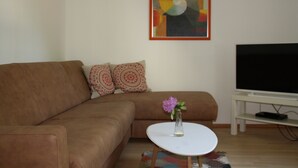 Un salon avec un canapé marron, une table basse blanche ornée d’un vase de fleurs, un téléviseur à écran plat posé sur un meuble et un tableau abstrait aux couleurs vives accroché au mur.